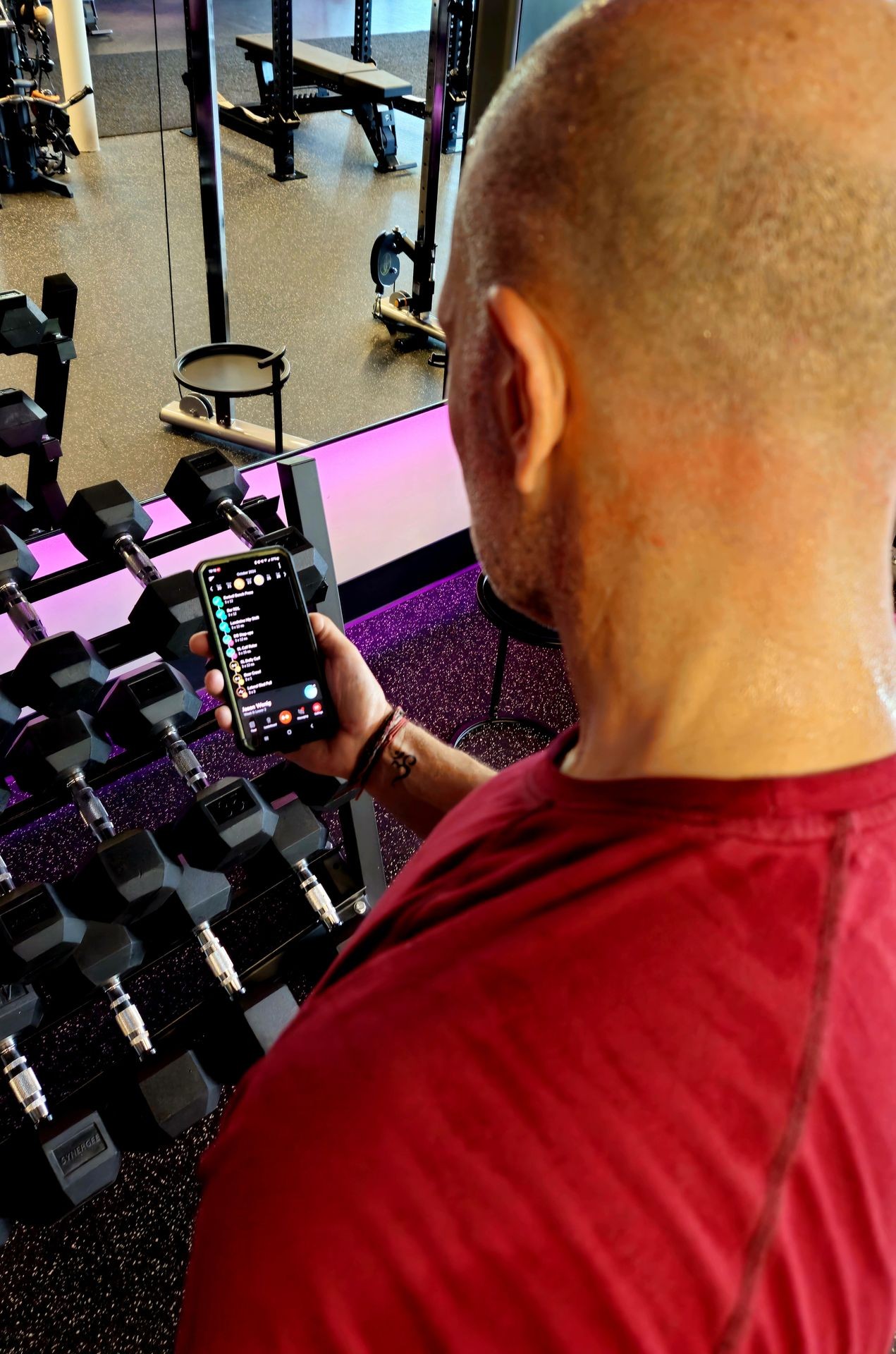 Person in a gym holding a smartphone near a rack of dumbbells. Online template programs by Rebecca Frey. Goal specific strength, fat loss, muscle gain, endurance plans delivered through her fitness app for independent clients seeking structured, flexible workouts without guesswork